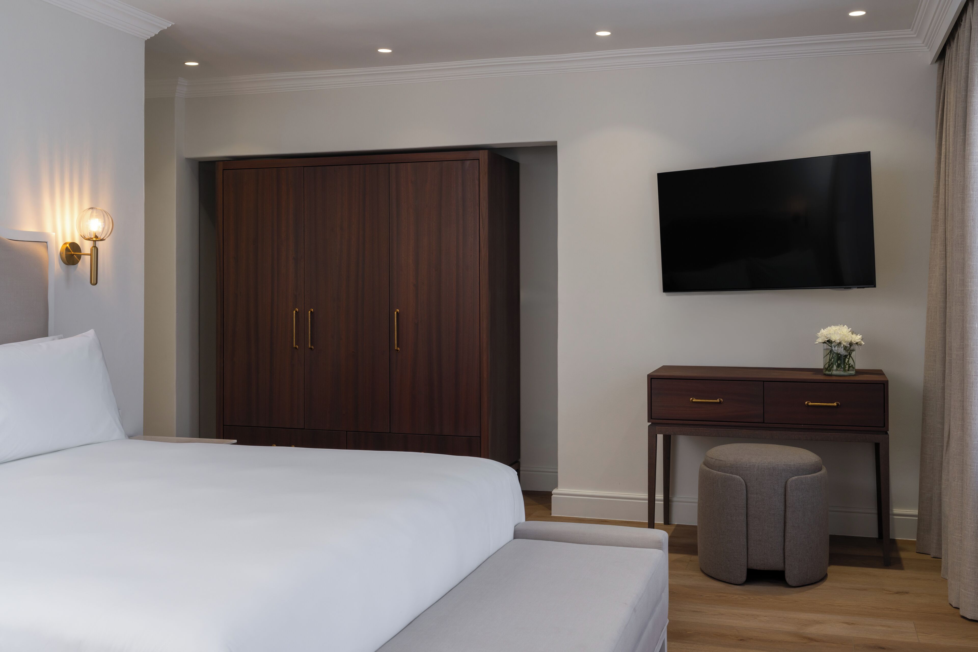A hotel room with a large bed, a wardrobe, a desk with drawers, a television mounted on the wall, and an ottoman.