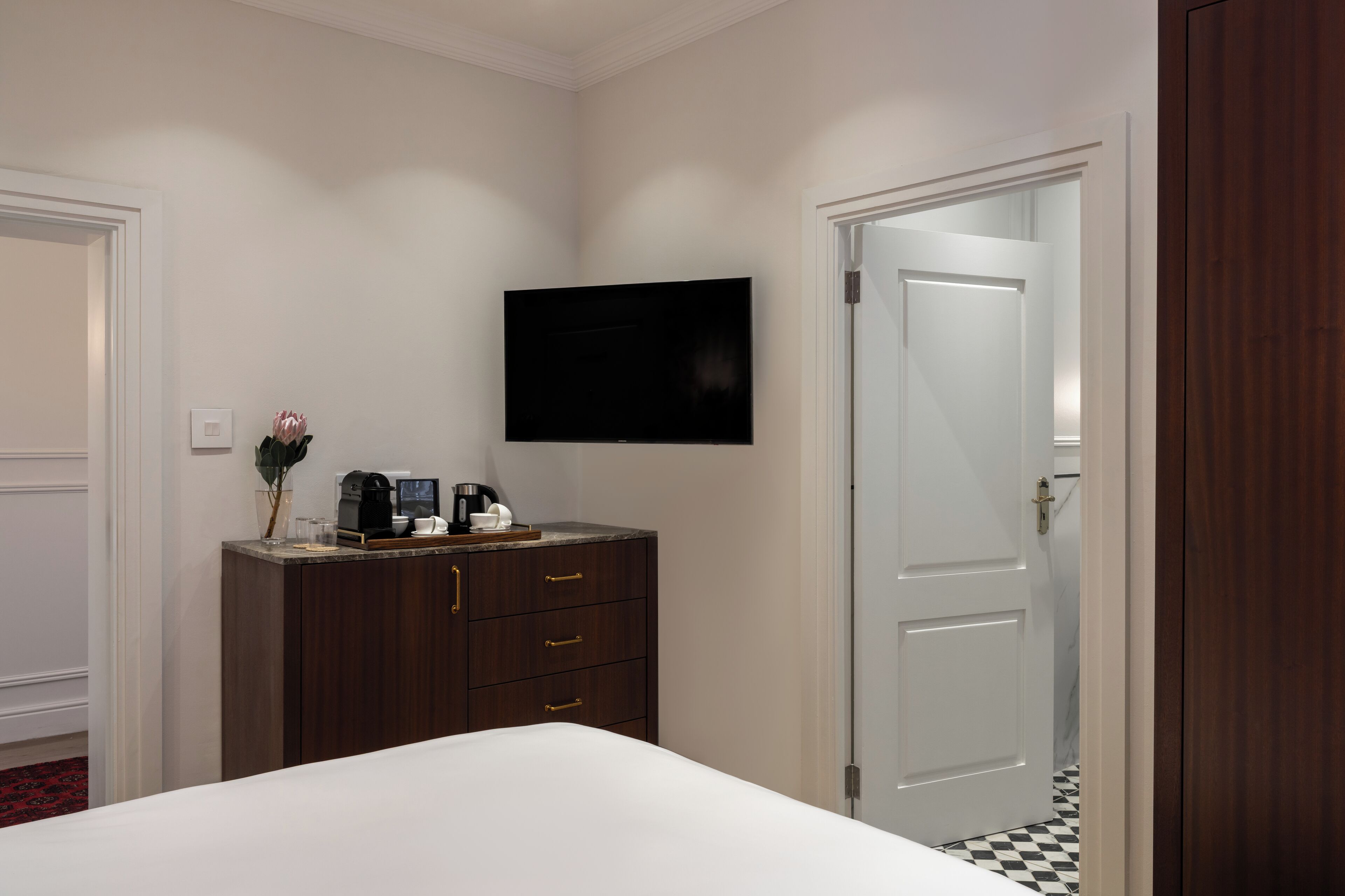 A hotel room with a bed, a dark wooden dresser, a flat-screen TV mounted on the wall, and a white door.