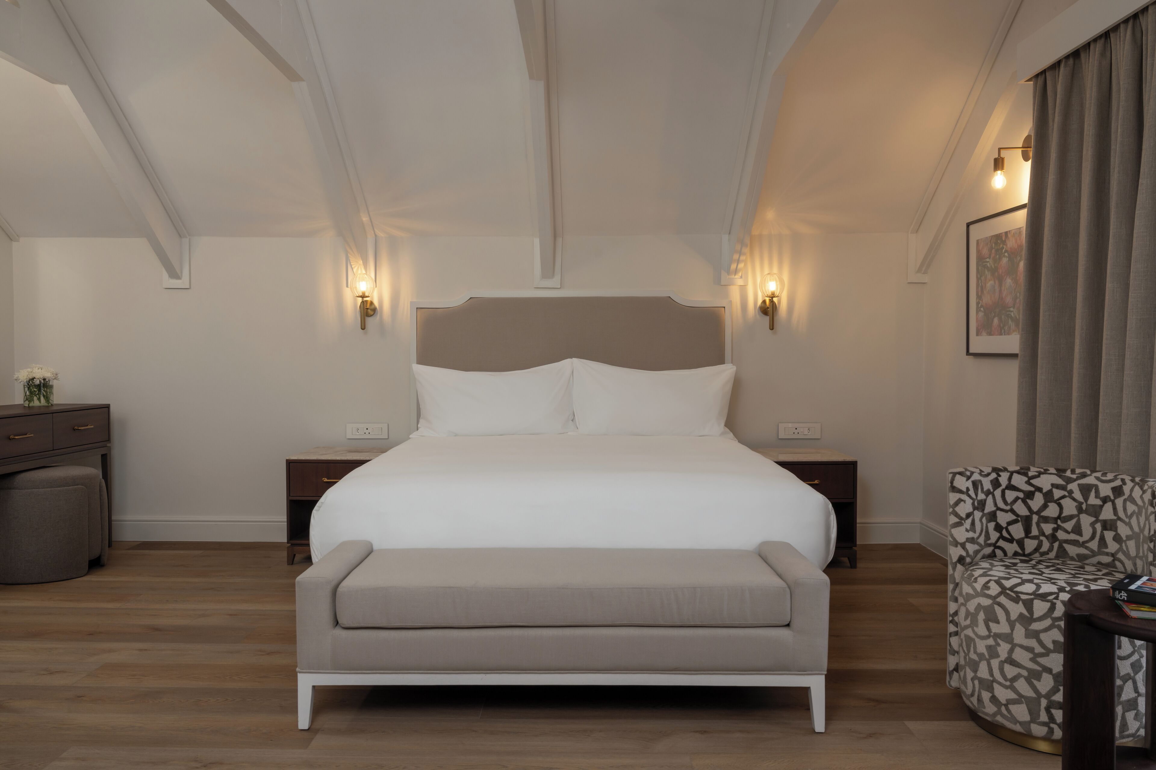 Un dormitorio con una cama grande, mesitas de noche, un banco, un sillón estampado y luces fijadas a la pared.
