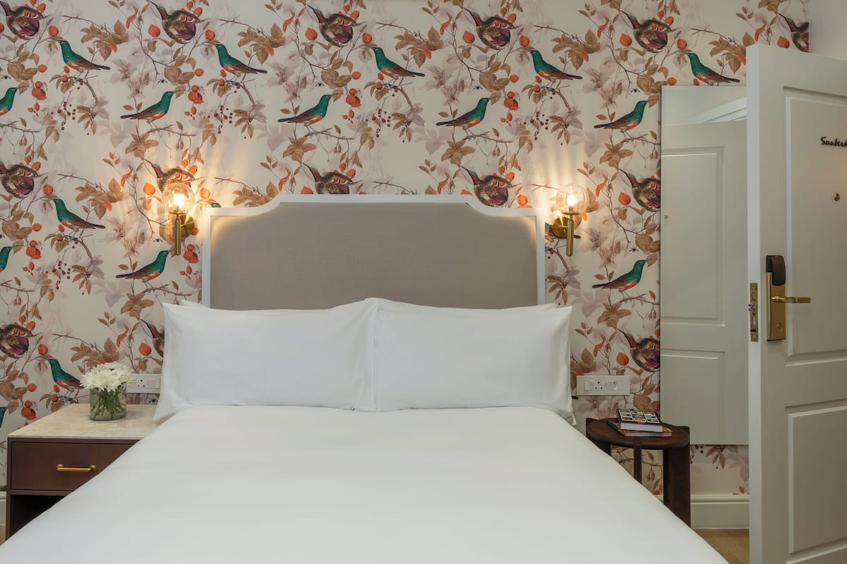 A neatly made bed with white linens, a wooden nightstand with a vase of flowers, and a wall with a floral wallpaper pattern.