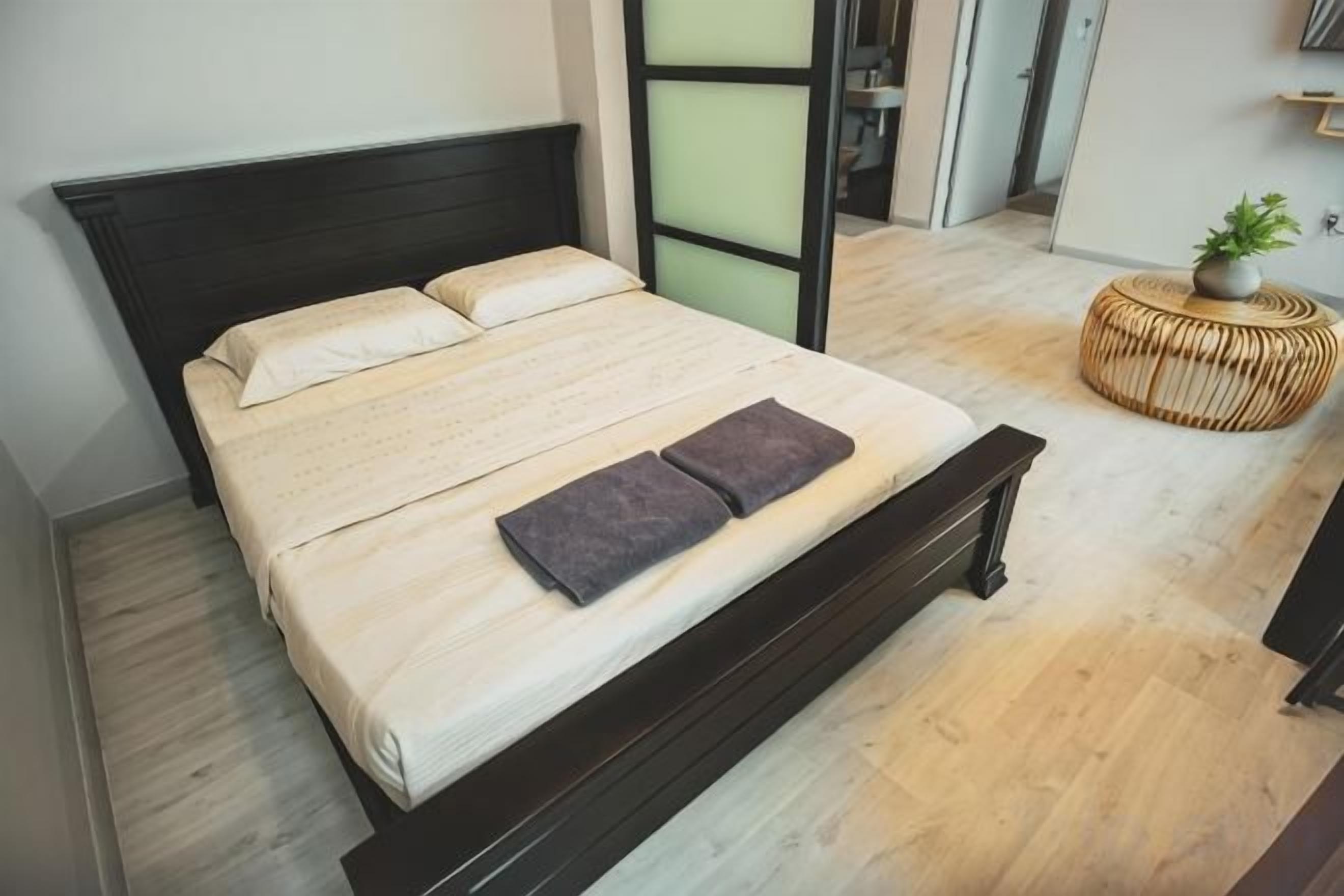Cama com cabeceira escura, colchão branco e duas almofadas. Pequena mesa redonda de vime com uma planta ao lado da cama.
