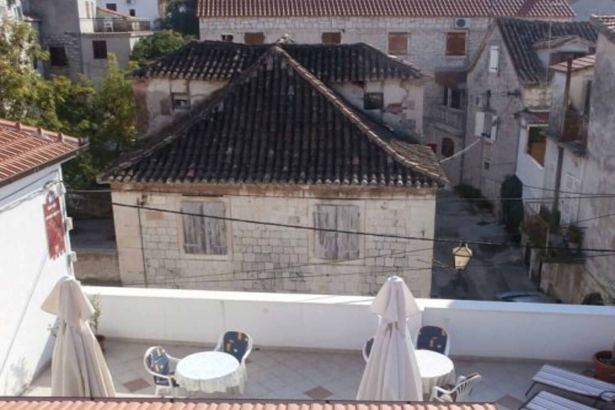 A rooftop terrace with a table and chairs, a small building with a tiled roof, and a narrow street below.