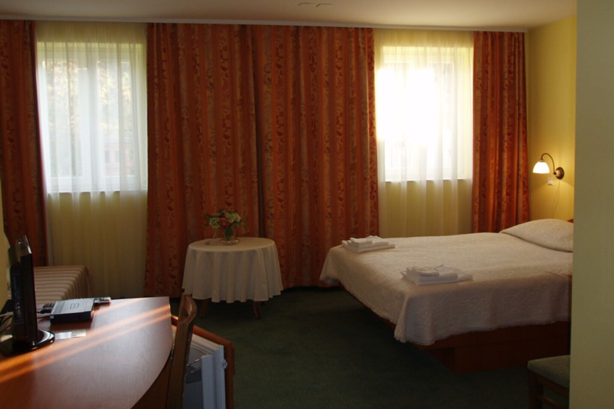 A hotel room with two beds, a desk, a small table with a vase, and a lamp.