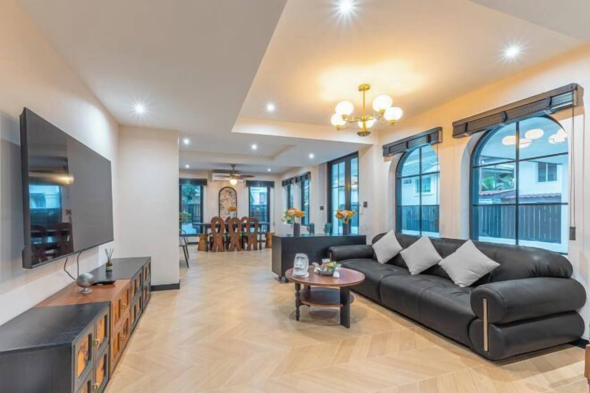 A spacious living area with a sofa, a coffee table, and a TV. There are large windows and a chandelier.