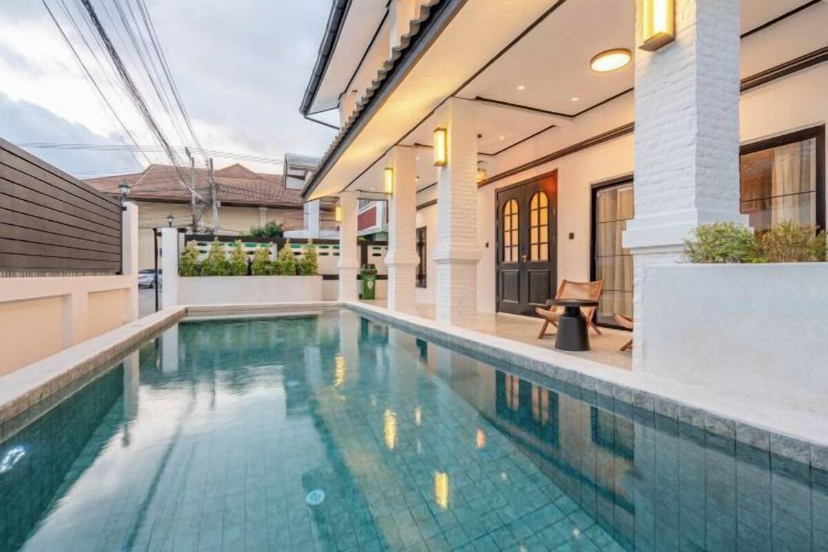 A swimming pool with a tiled deck, a covered area with seating, and a building with a traditional architectural style.