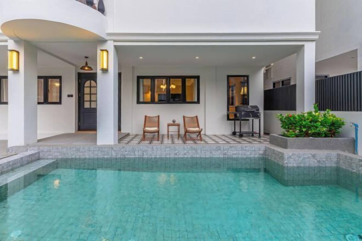 A pool area with a clear pool, two chairs, a grill, and a potted plant.