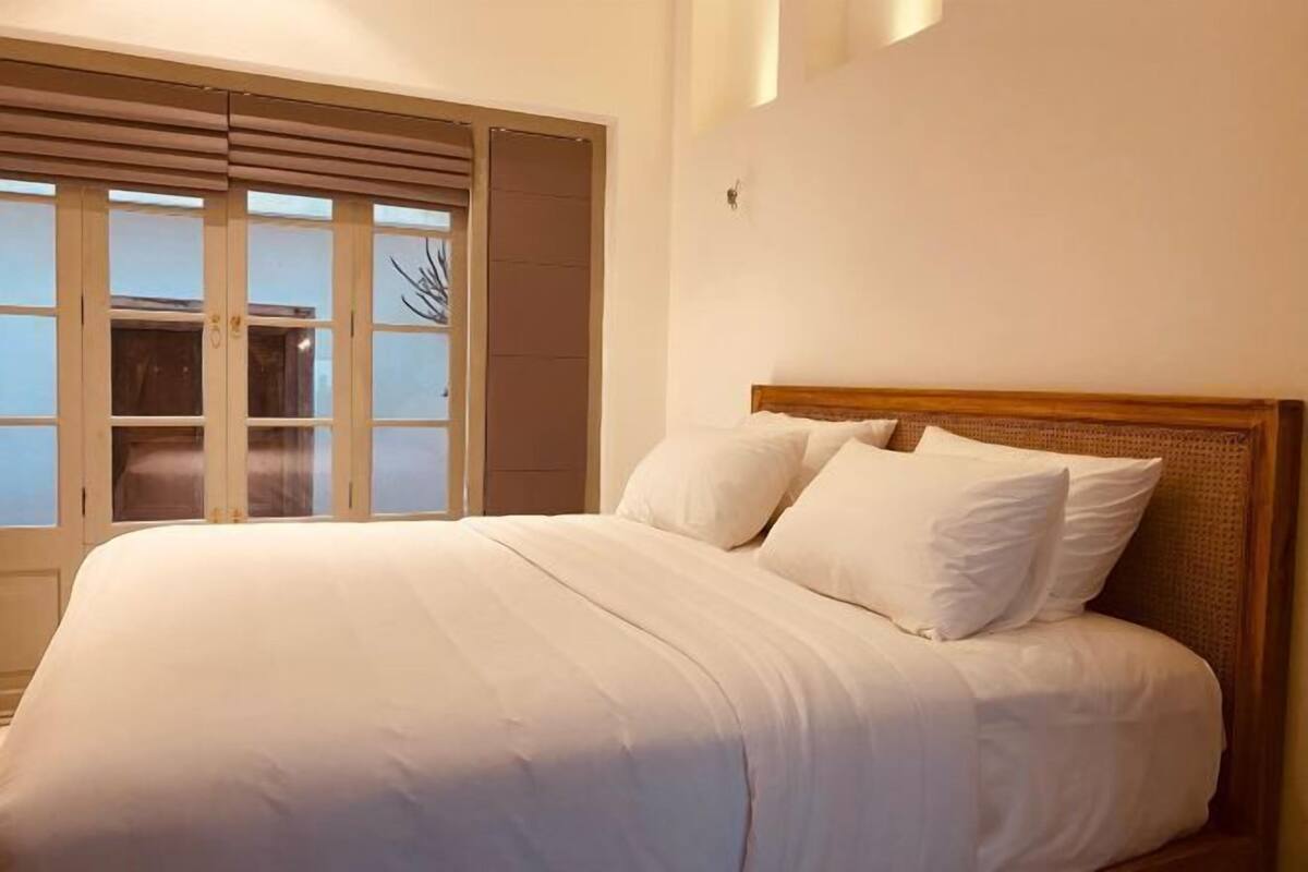Een keurig opgemaakt bed met witte lakens in een kamer met een groot raam en een houten hoofdbord.