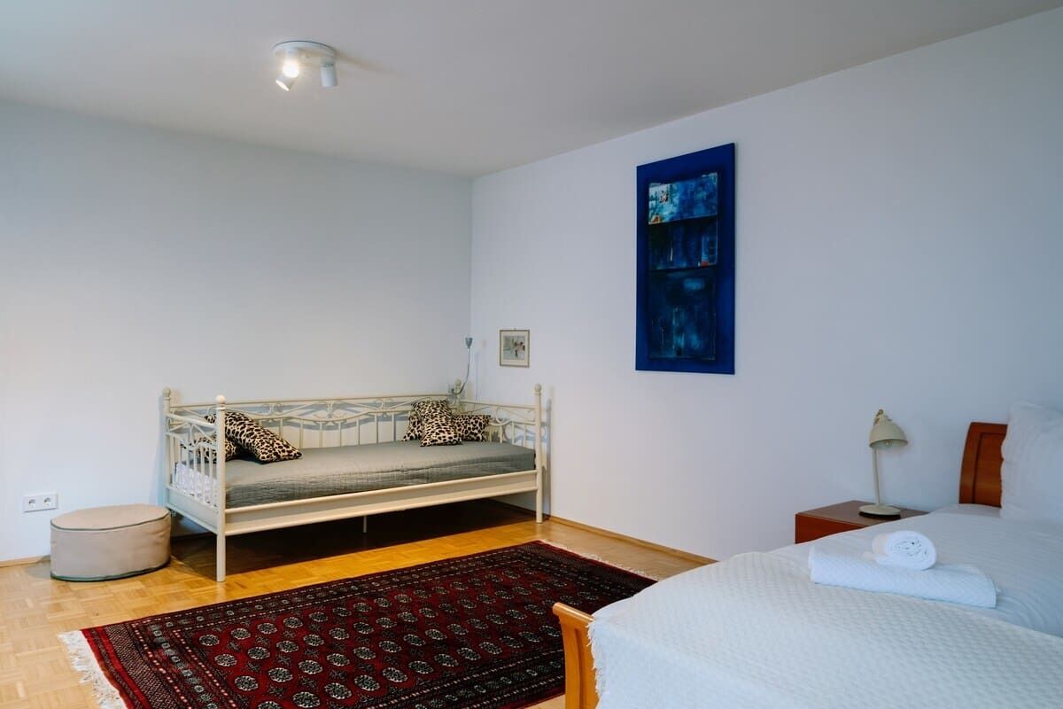 Ein Schlafzimmer mit einem Bett, einer Schlafcouch, einem kleinen runden Hocker, einem roten Teppich mit Muster, einem Nachttisch mit Lampe und einem gerahmten Bild an der Wand.