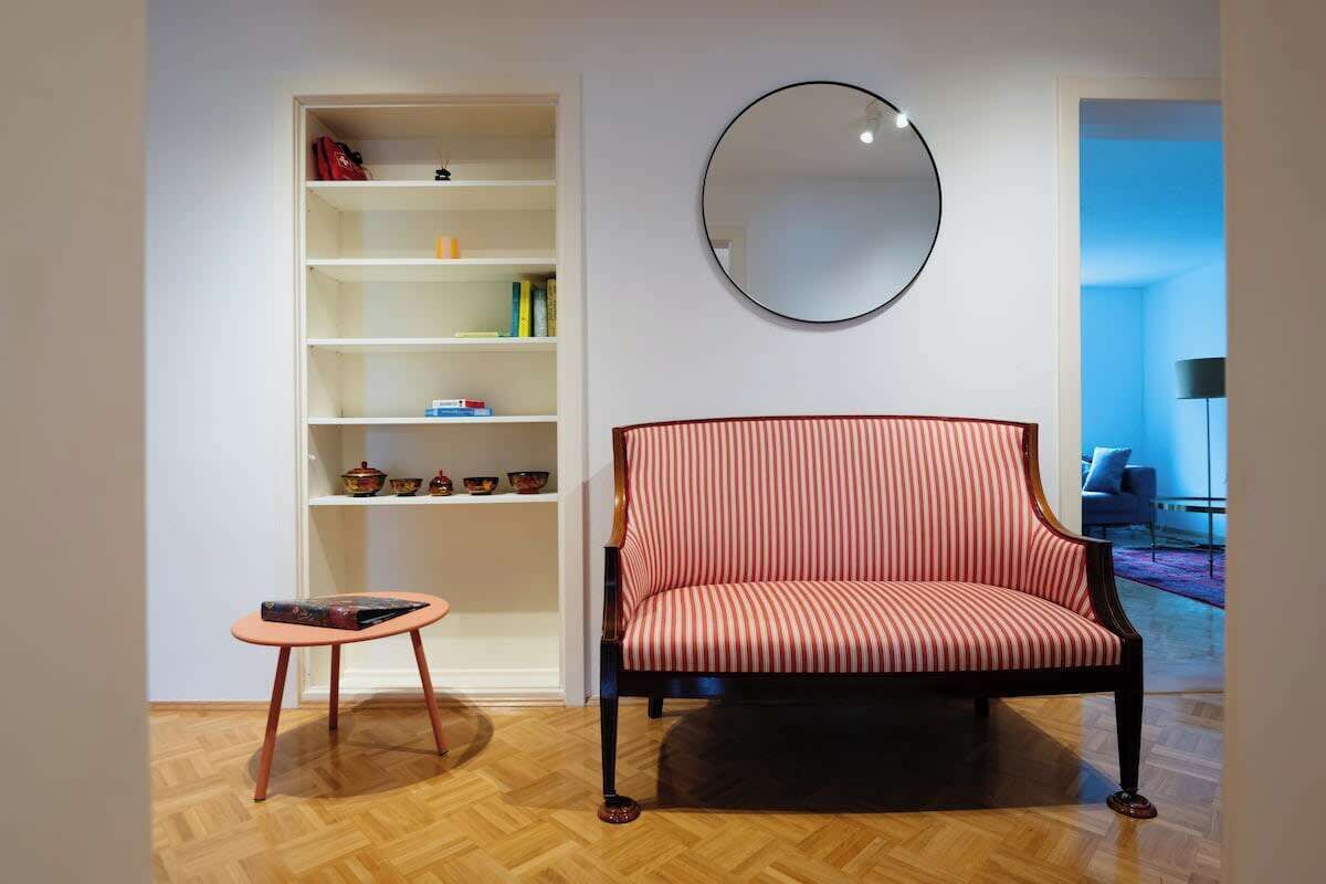 Een kamer met een gestreept fauteuil, een klein tafeltje, een boekenkast en een ronde spiegel aan de muur.