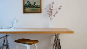 A desk with a white lamp, a vase with dried flowers, and a painting on the wall.