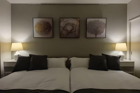 Una cama doble con sábanas blancas, dos almohadas negras y una mesita de noche con lámpara a cada lado.
