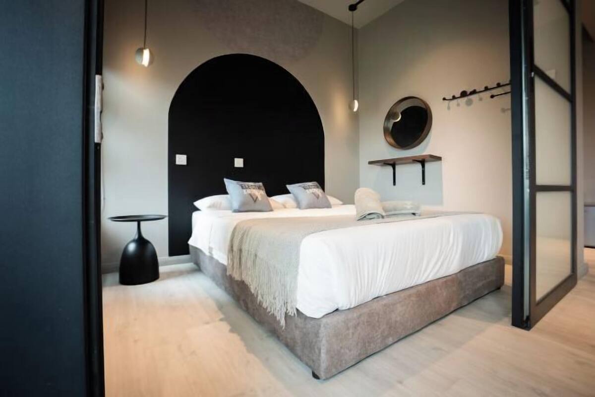 Un dormitorio moderno con una cama grande, un espejo redondo, una mesita pequeña y un arco negro original.