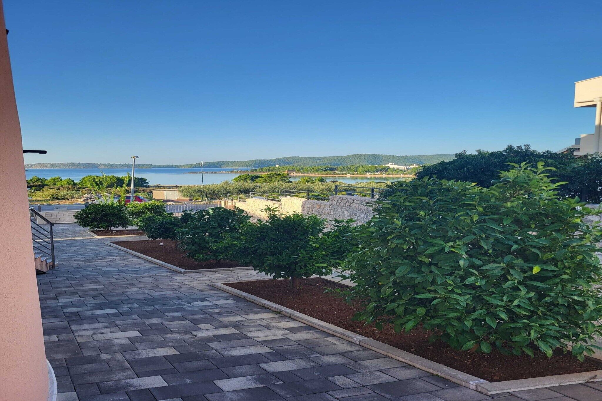 A paved pathway leading to a balcony overlooking a body of water, with greenery and a distant view of hills.