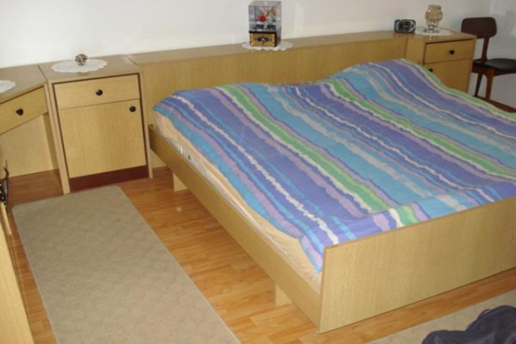 A single bed with a colorful striped bedspread, a wooden nightstand with a small lamp, and a chair in a simple room with wooden flooring.