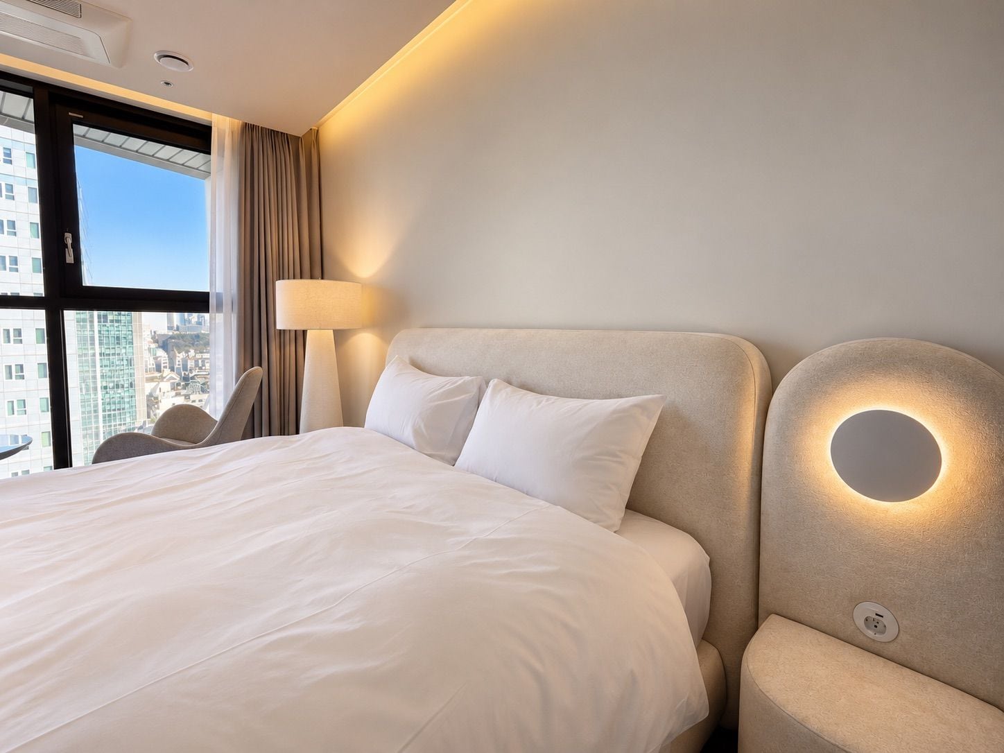 A hotel room with a large bed, a bedside lamp, a chair, and a view of the city through the window.