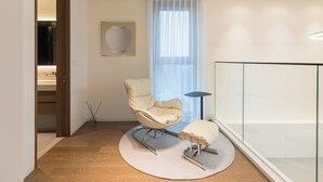 Een moderne woonkamer met een rond tapijt, een gestoffeerde kruk, een fauteuil, een vloerlamp en een groot raam met dunne gordijnen.