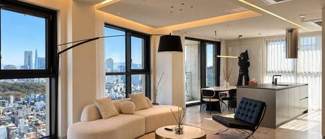 Un salon moderne comprenant un canapé, un pouf et une table basse, de grandes fenêtres offrant une vue sur la ville, ainsi qu’un coin repas avec un îlot de cuisine.