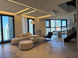 Ein modernes Wohnzimmer mit Sofa, Hocker und Sessel, ein Essbereich mit Tisch und Stühlen, große Fenster mit Blick auf die Stadt und eine Treppe.