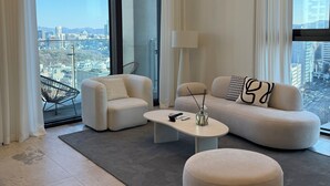 Un salon moderne comprenant un canapé, un fauteuil, un pouf et une table basse. De grandes fenêtres offrent une vue sur la ville.