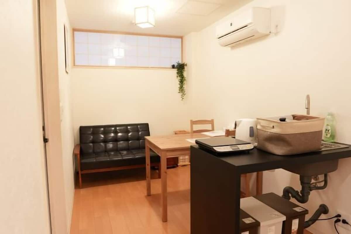 Una habitación pequeña con un sofá de cuero negro, una mesa de madera, una ventana con vistas, aire acondicionado y un escritorio con una bolsa y una botella.