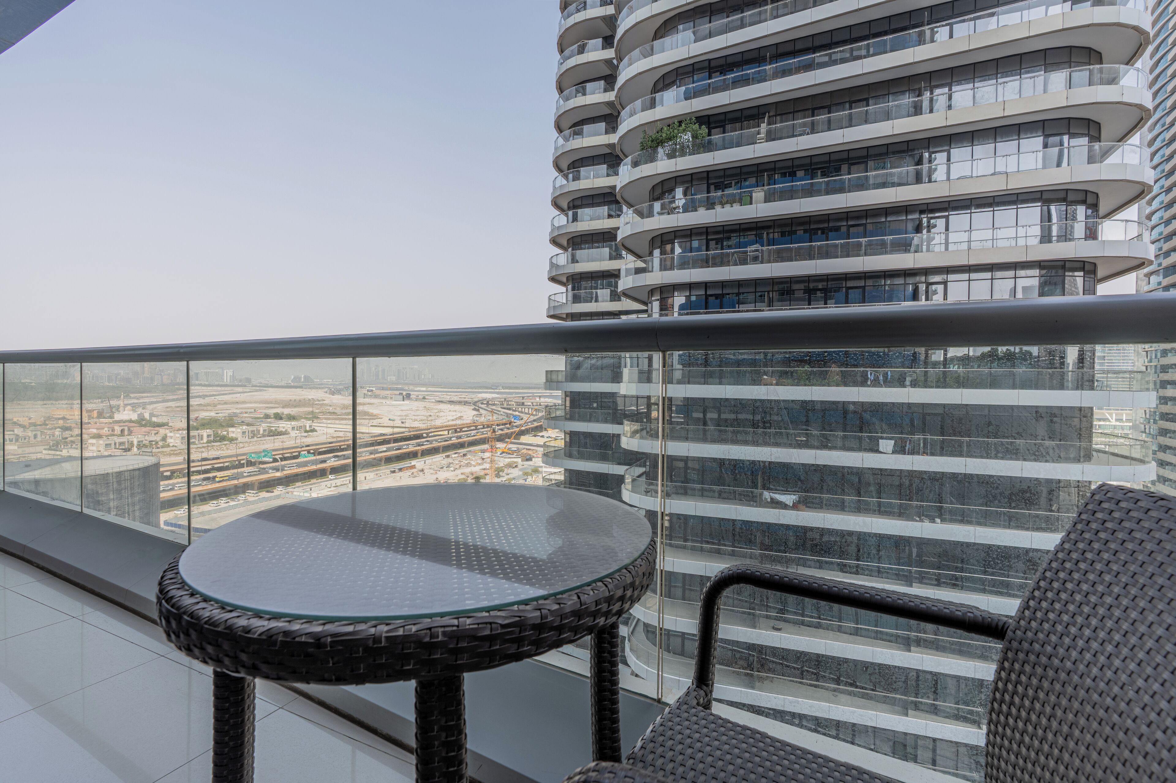 Un balcon moderne avec une table en verre et des chaises en osier, offrant une vue sur un paysage urbain avec un immeuble de grande hauteur et une voie ferrée.