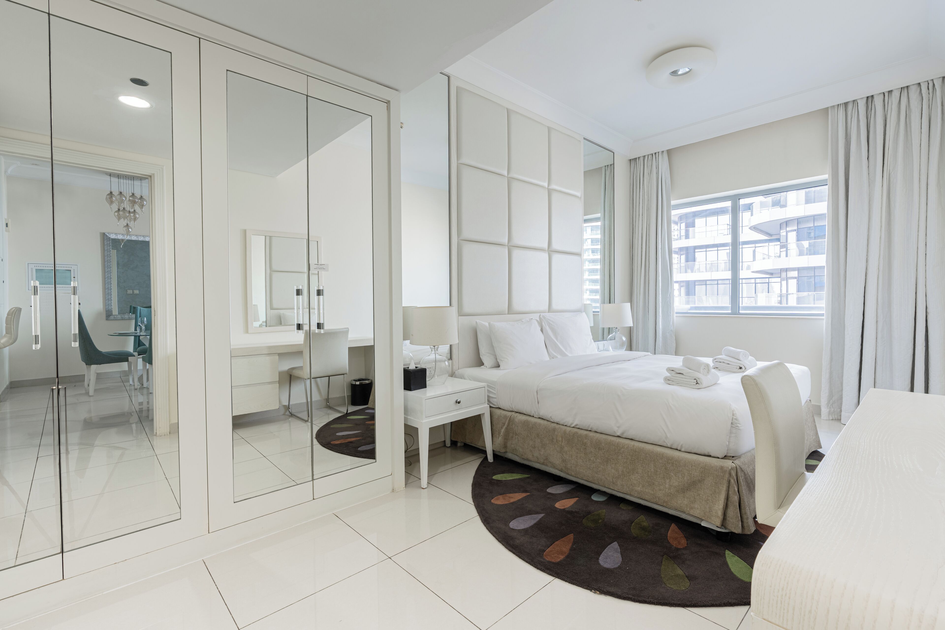 A modern hotel room with a large bed, a desk, a chair, and a view of the city through the window.
