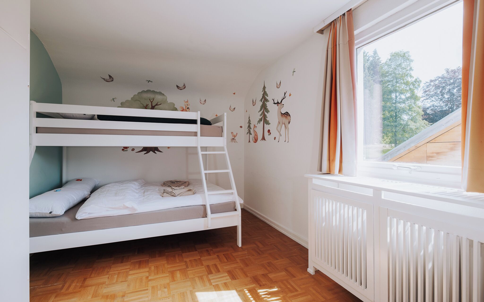 A bunk bed room with a wooden floor, a window with curtains, and wall decals of animals and trees.