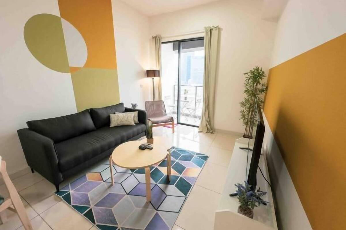 Een moderne woonkamer met een zwarte bank, een ronde salontafel, een tapijtend met een patroon, een televisie en een balkon met gordijnen.