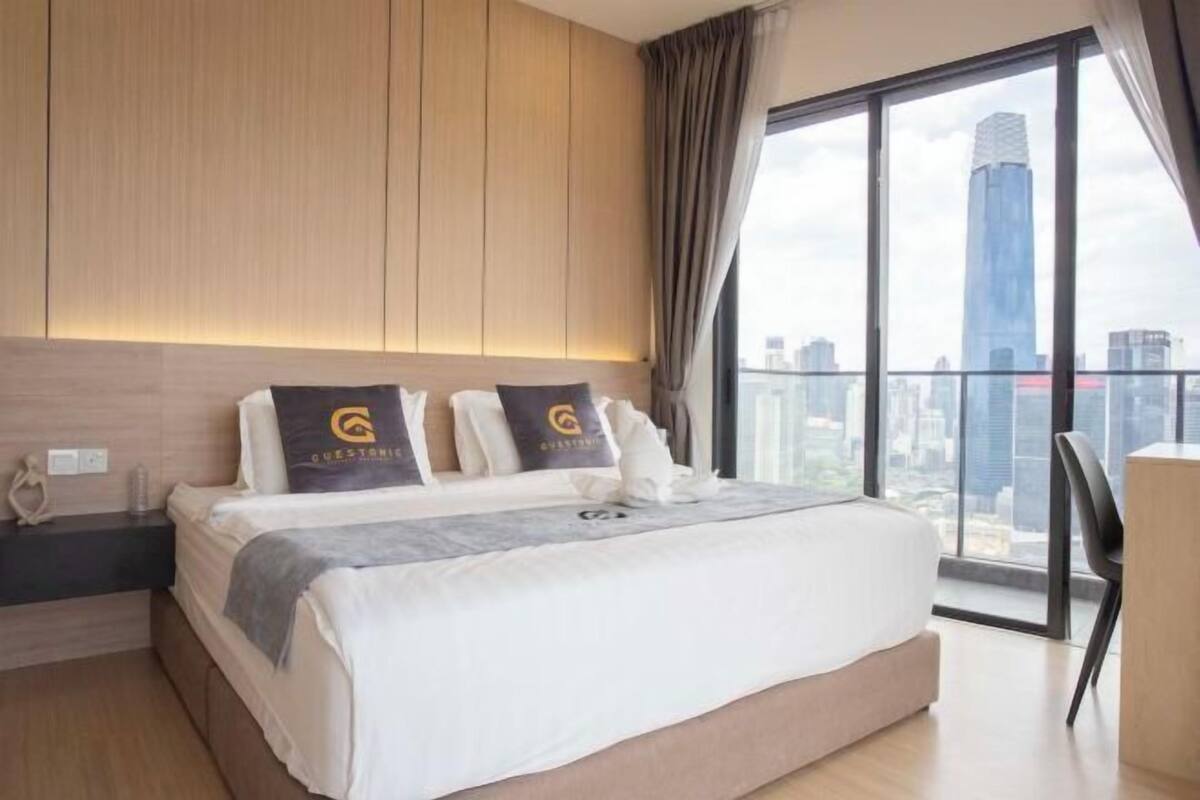 Een moderne hotelkamer met een groot bed, een nachtkastje, een bureau en uitzicht op een stadsbeeld door grote ramen.