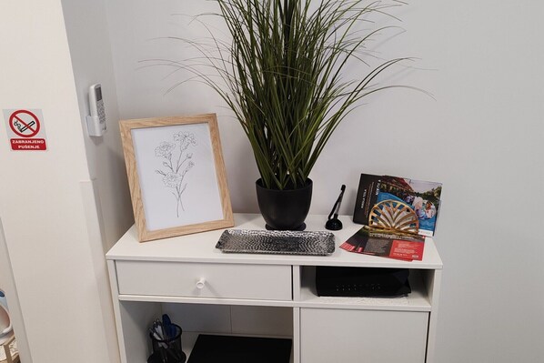 Un escritorio blanco con una ilustración botánica enmarcada, una planta en maceta, un plato decorativo y un folleto.
