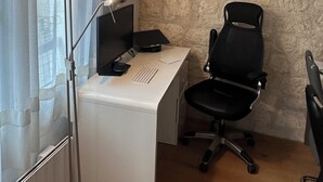 Ein kleiner Büroraum mit einem weißen Schreibtisch, einem schwarzen Bürostuhl, einer Stehlampe und einer Steinwand.
