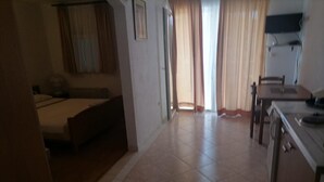 A hotel room with a bed, a desk, and a chair. There is a window with curtains, and a door leading to another room. - Apartments Libertas - Studio with Balcony and Sea View - (3) (Rogoznica)