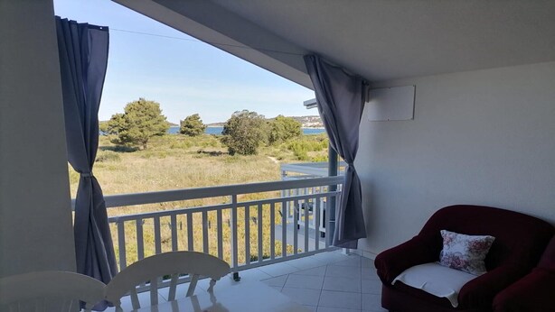 A balcony with a white table and chairs, a red sofa, and a view of a grassy area and trees. - Apartments Libertas - Two Bedroom Apartment with with Terrace and Sea View (7) (Rogoznica)