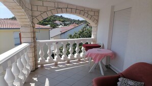 Un balcón con barandilla blanca, un sofá rojo, una mesita con mantel rosa y vistas a un río y edificios al fondo.
