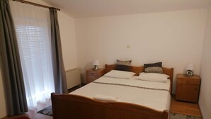 Una cama bien hecha con cabecera de madera, dos mesitas de noche con lámparas, una silla roja y una ventana con cortinas traslúcidas.