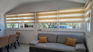 Una sala moderna con un sofá gris, dos cojines color mostaza, una mesa de comedor con sillas y ventanas amplias con persianas horizontales.