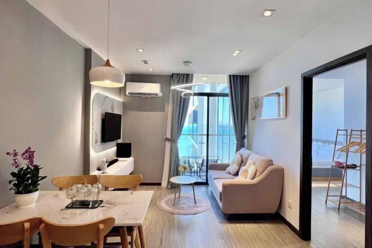 A modern living room with a sofa, dining table, and chairs, a TV mounted on the wall, and a large window with curtains.