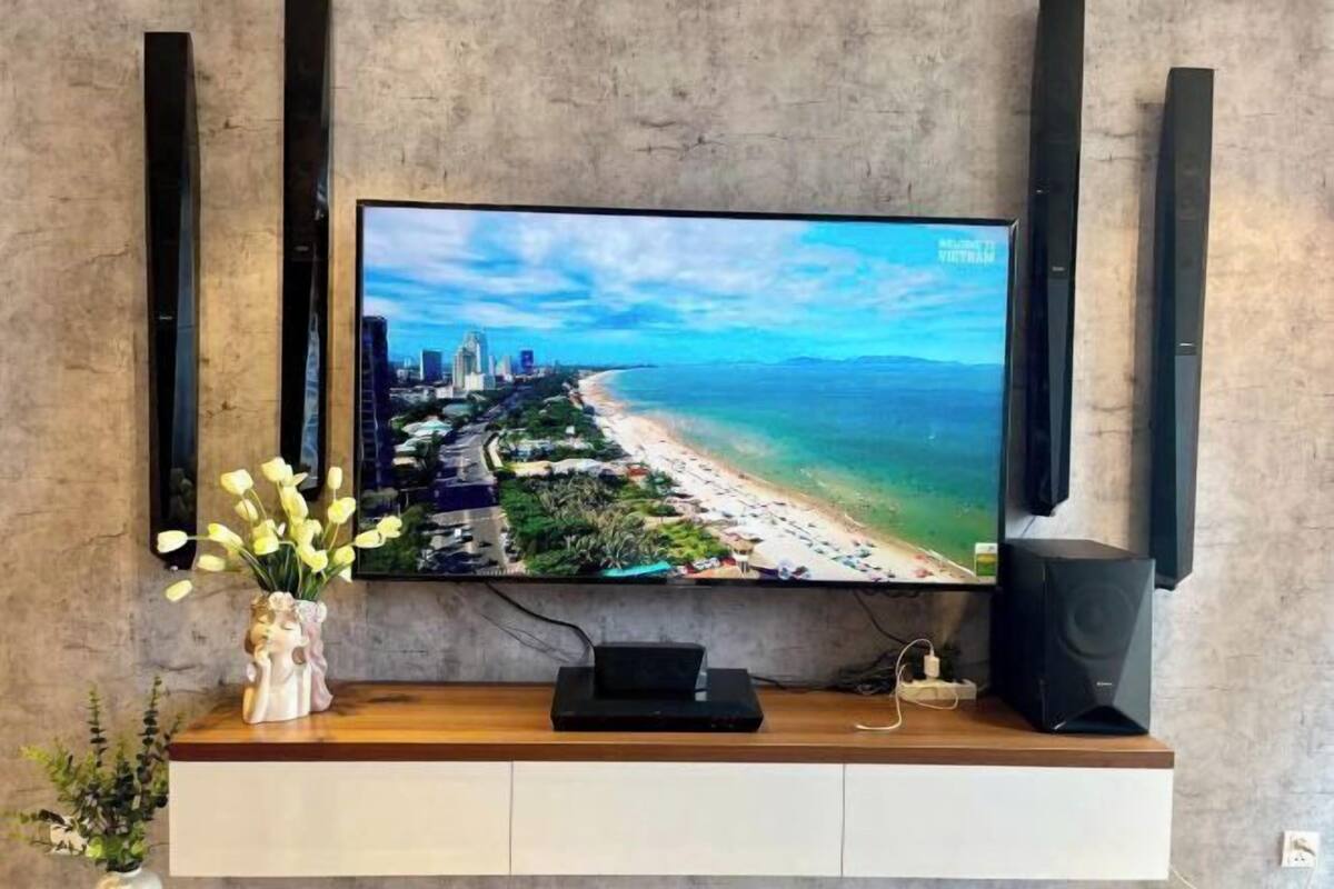 A flat-screen TV mounted on a wall with a wooden console below, a small black speaker, and a vase with flowers.