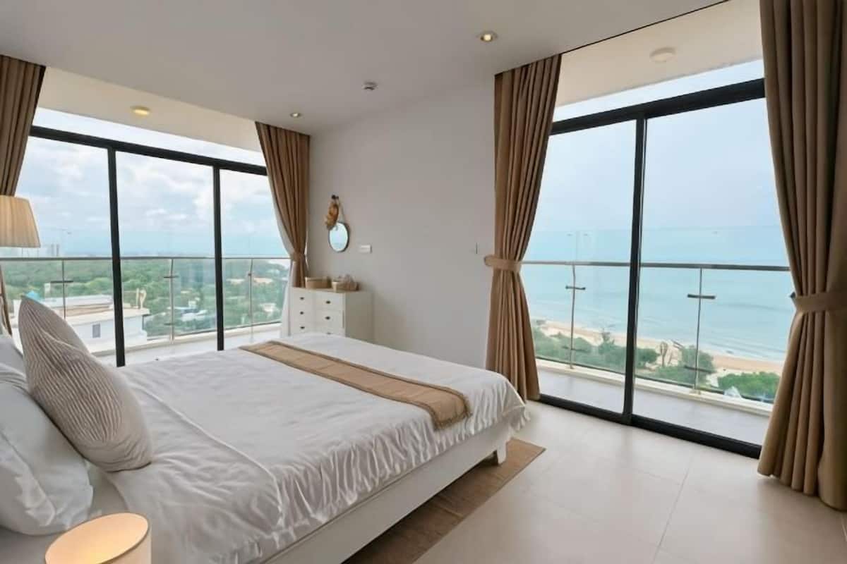 A bedroom with a large bed, a dresser, and a view of the ocean through floor-to-ceiling windows.