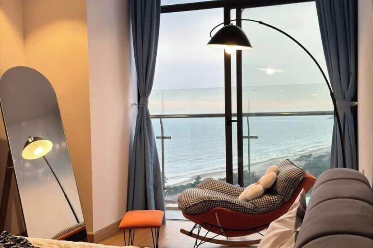 A modern room with a large window, a rocking chair, a small stool, and a floor lamp.