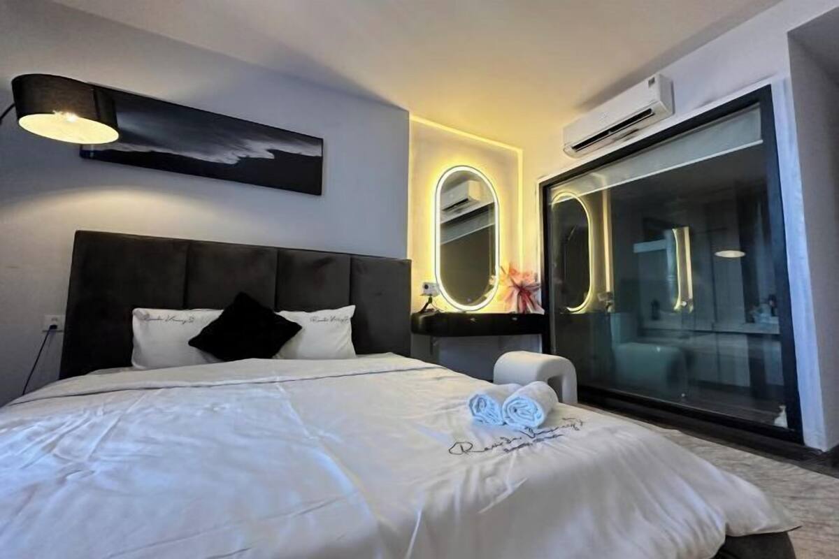 A modern hotel room with a large bed, a bedside table with a lamp, a mirror, and a sliding glass door leading to a bathroom.