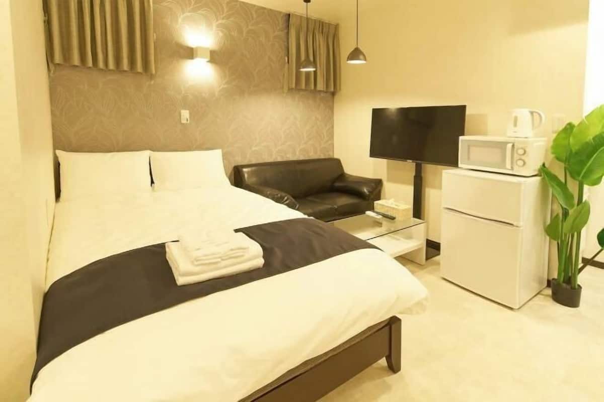 Habitación de hotel con una cama, un sillón de cuero negro, un escritorio con televisor de pantalla plana, un microondas y una planta en maceta.