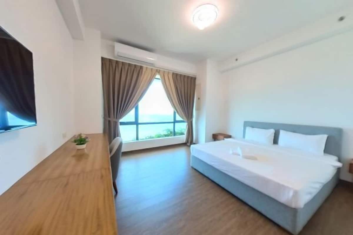 A hotel room with a bed, a wooden table, a small plant, a window with curtains, and a ceiling light.