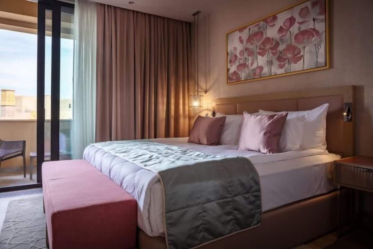 Ein Hotelzimmer mit Bett, Sitzbank, Nachttisch, Wandlampe und einem Fenster mit Vorhängen.