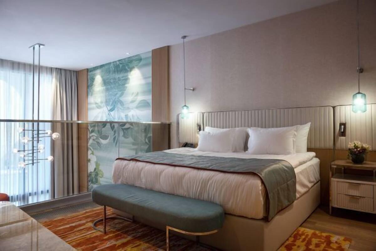 Ein modernes Hotelzimmer mit einem großen Bett, einer Bank, einem Nachttisch und Blick auf eine Stadtlandschaft durch raumhohe Fenster.