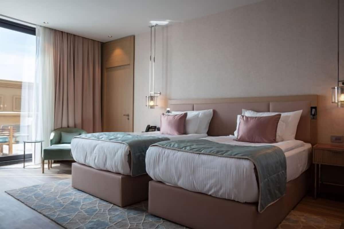 Ein Hotelzimmer mit zwei Betten, einem Sessel, einem Schreibtisch und einem großen Fenster.