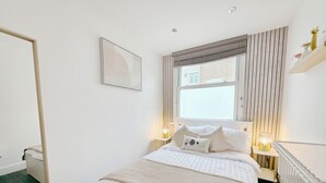 Ein Schlafzimmer mit einem Bett, Nachttischlampen, einem Fenster mit Jalousien, einem Spiegel und einem Wandbild.