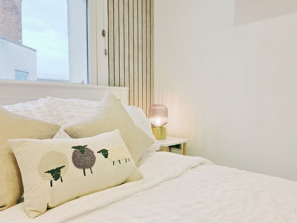 A neatly made bed with white bedding and decorative pillows, a bedside table with a lamp, and a window with blinds.