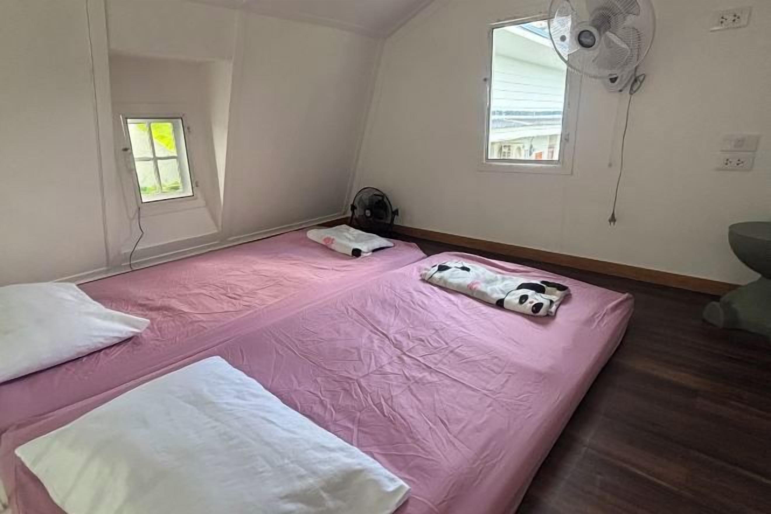 A bedroom with a pink bedspread, two pillows, a fan, and a window.