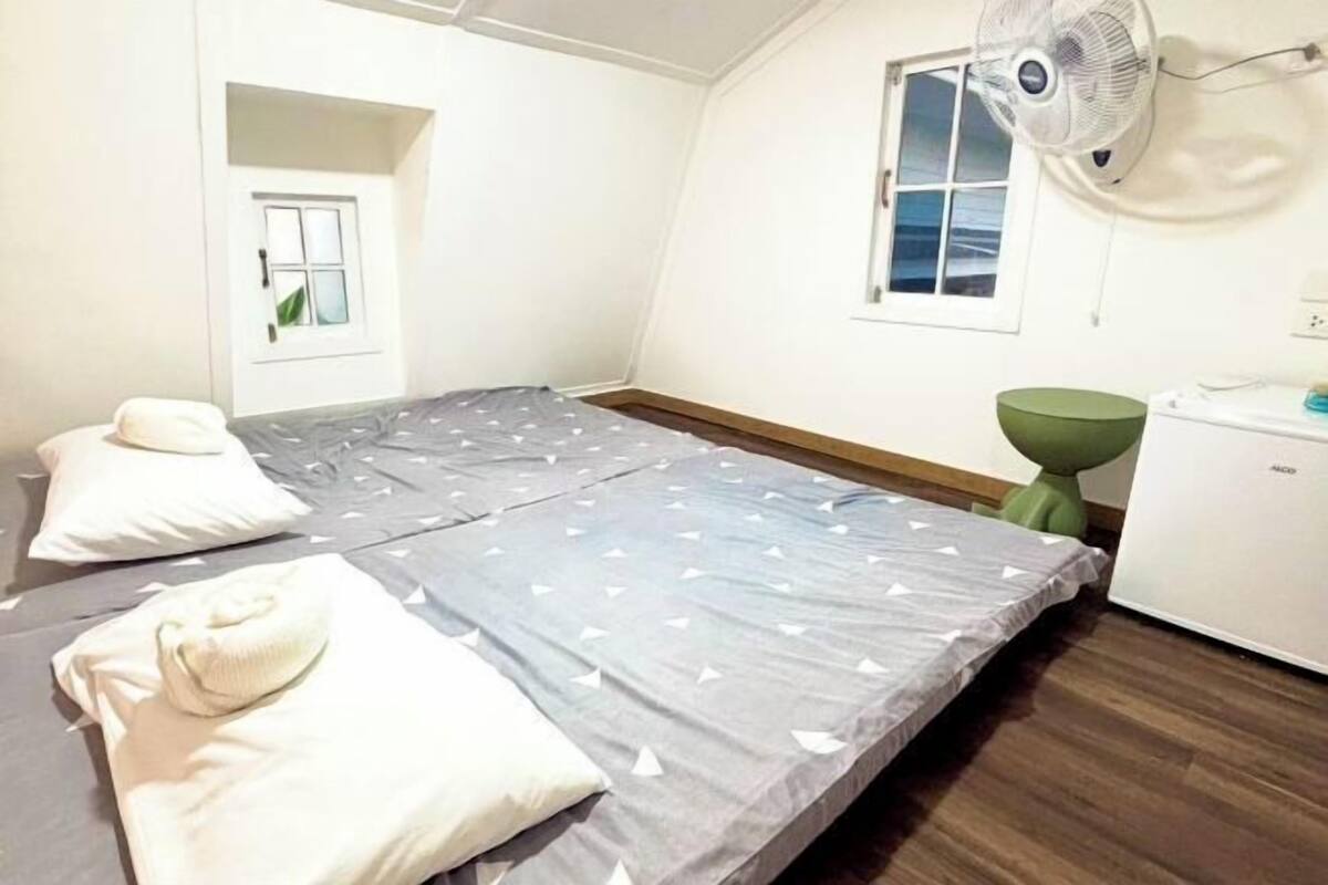 A small, single-bed room with a fan, a small table, and a window.
